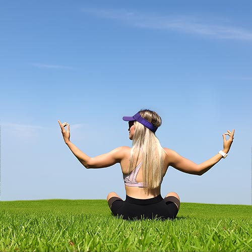 A woman with long blonde hair sitting cross-legged on green grass, meditating under a clear blue sky, viewed from behind