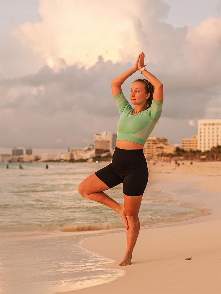 A woman practicing yoga in tree pose on a sandy beach at sunset, with ocean waves and city buildings in the background